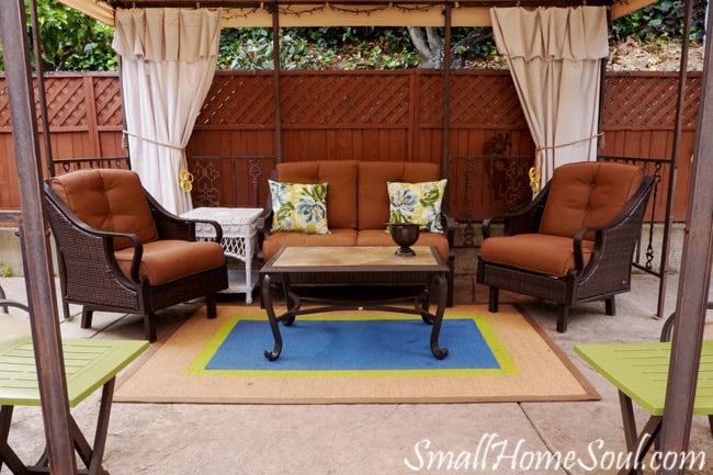 Drop cloth curtains on patio at back of gazebo with patio furniture placed around patio rug.