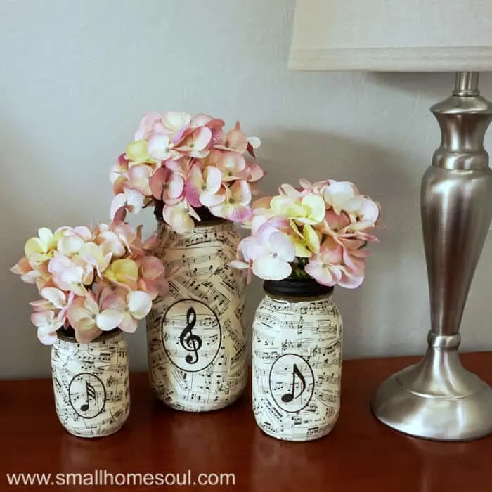 Three sheet music mason jars on table filled with hydrangeas next to lamp.