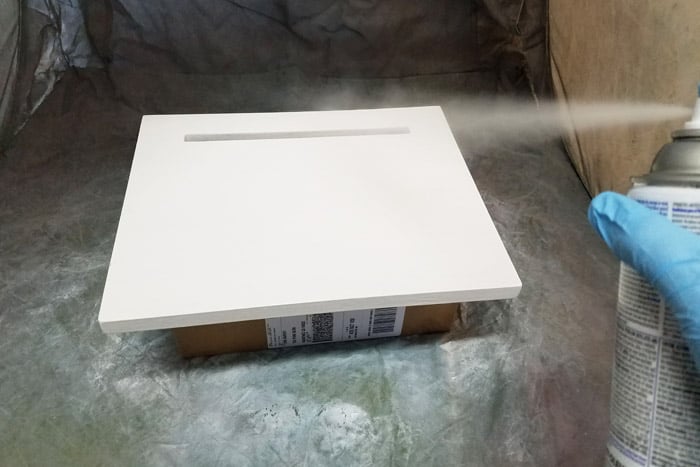 Spray painting lid of box white while propped on box in small spray shelter.