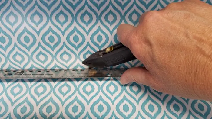 hand holding utility knife cutting contact paper against metal ruler.