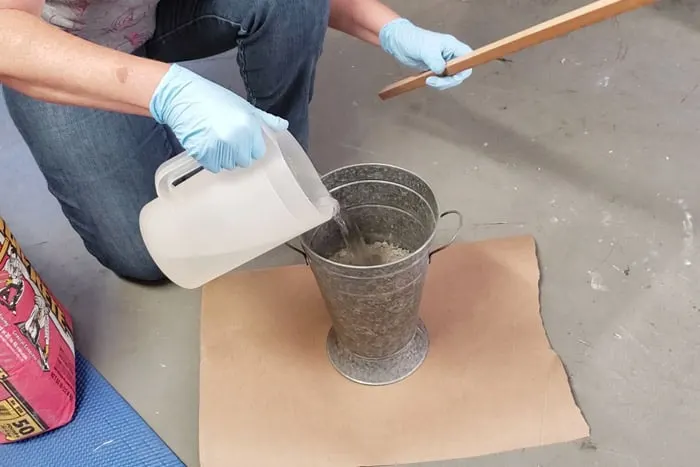 gloved hands pouring water from a pitcher into galvanized bucket