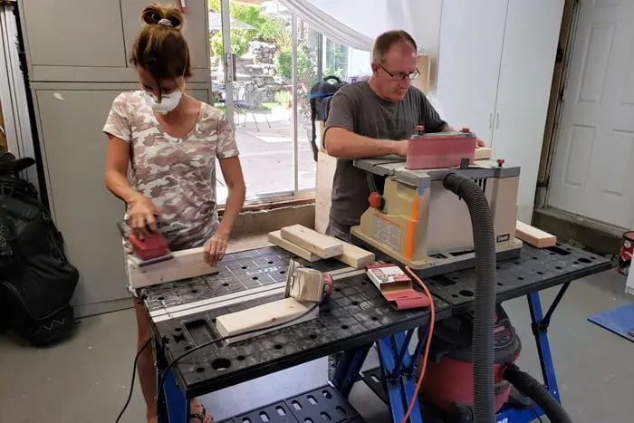 Man and woman using power sanders to sand 2x4s at work bench.