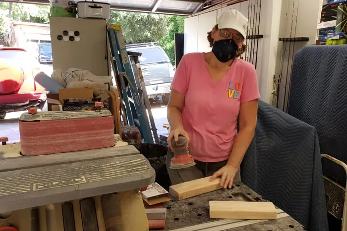 Woman in pink shirt and white hat wearing black dust mask sanding 2x4s in garage.