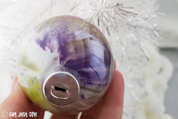 Green and purple paint pour christmas ornament in front of white tree.