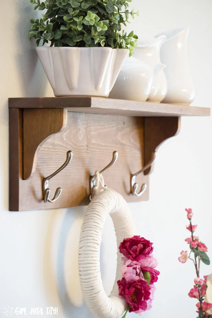 Wooden coat rack attached to wall with wreath hanging from center hook and white vases on the shelf.