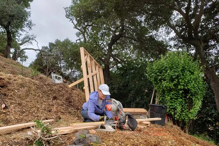 Woman crouched at saw on hillside path cutting wood.