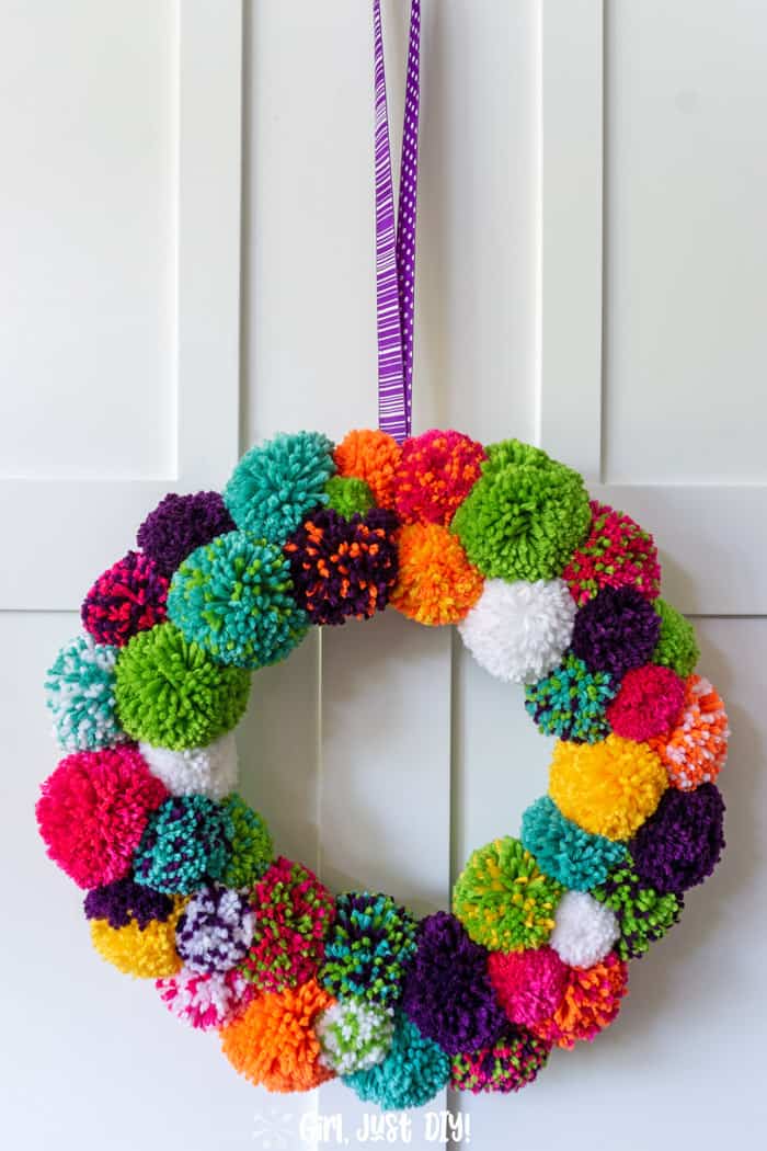 Tall view of diy pom pom wreath hanging on white door from purple ribbon.