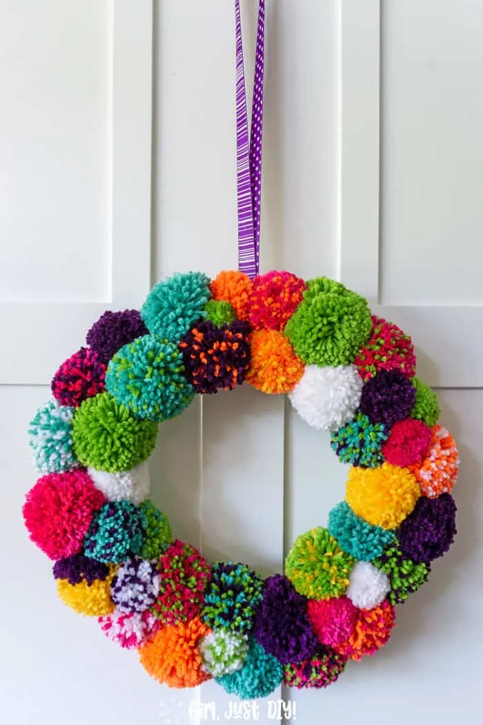 Tall view of diy pom pom wreath hanging on white door from purple ribbon.
