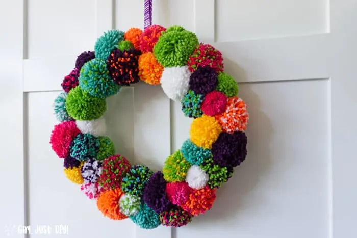Wide picture of completed DY Pompom Wreath hanging on white door from purple ribbon.