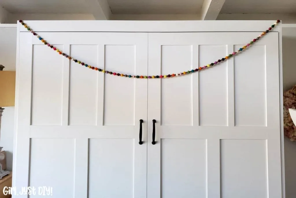 Colorful felt ball garland hanging across white cabinet face.